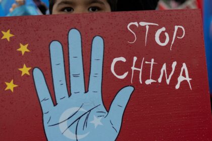Grupo de uigures protestando con carteles contra la represión china en Xinjiang, rodeados de alambradas y vigilancia