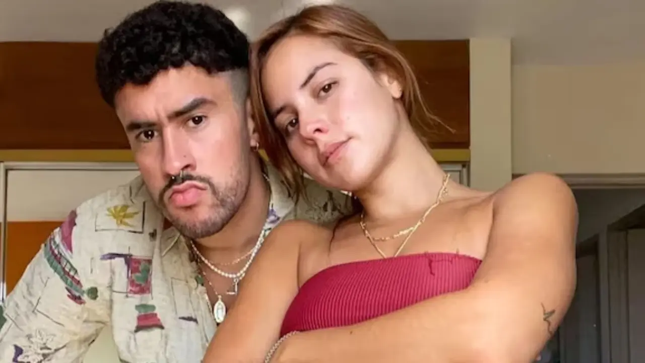 Bad Bunny y Gabriela Berlingeri sonriendo en una playa australiana durante la gira Debí Tirar Más Fotos, con gafas de sol y ropa casual.