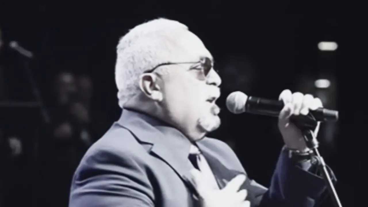 Willie Colón tocando el trombón en un escenario, rodeado de músicos, con expresión intensa y pasión por la salsa