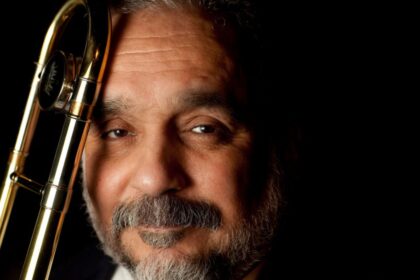 Willie Colón en un escenario con traje oscuro, rodeado de músicos, mientras sostiene una trompeta, reflejando su legado musical y las controversias que lo acompañaron