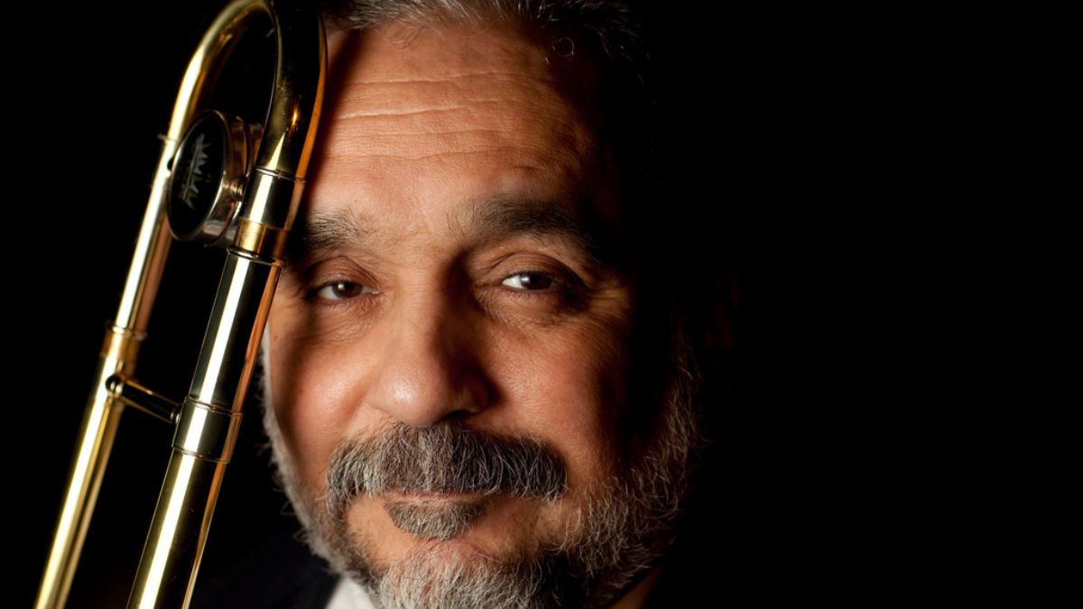 Willie Colón en un escenario con traje oscuro, rodeado de músicos, mientras sostiene una trompeta, reflejando su legado musical y las controversias que lo acompañaron