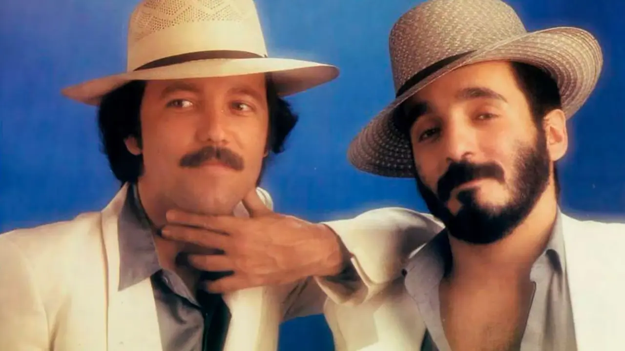Rubén Blades y Willie Colón en un escenario oscuro, iluminados por un foco, con partituras de 'El Baquiné de los Angelitos Negros' al fondo