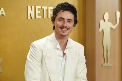 Timothée Chalamet con gesto incómodo en debate mientras un cartel de 'Akenatón - Ópera de Los Ángeles' brilla al fondo