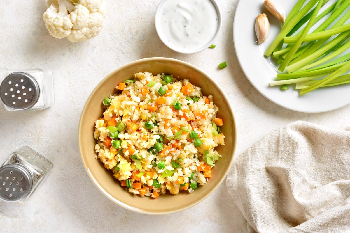 Plato de arroz frito de coliflor con camarones, zanahoria rallada y cebollín, servido en un bol de cerámica blanca con palillos al lado