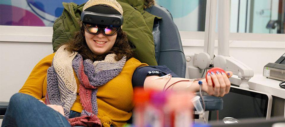 Mujer usando visor de realidad mixta durante donación de sangre, con animación inmersiva de paisaje relajante en pantalla 4K