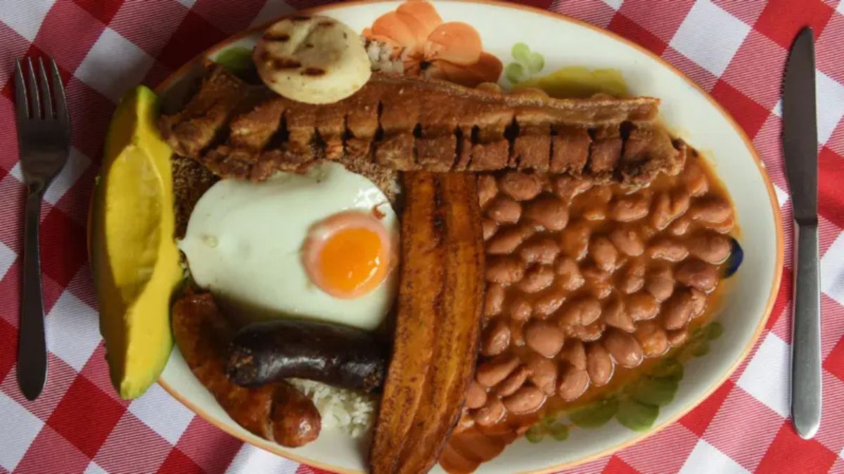 Bandeja paisa completa con arroz, frijoles, chicharrón, huevo, plátano, aguacate y arepa sobre mesa rústica, símbolo de la gastronomía colombiana que mezcla tradición y controversia