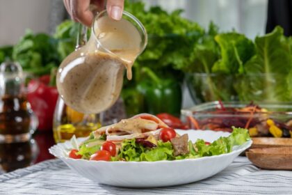 Cinco aderezos caseros en frascos de vidrio sobre mesa con ingredientes frescos: aguacate, miel, tahini, limones y hierbas aromáticas para ensaladas vibrantes