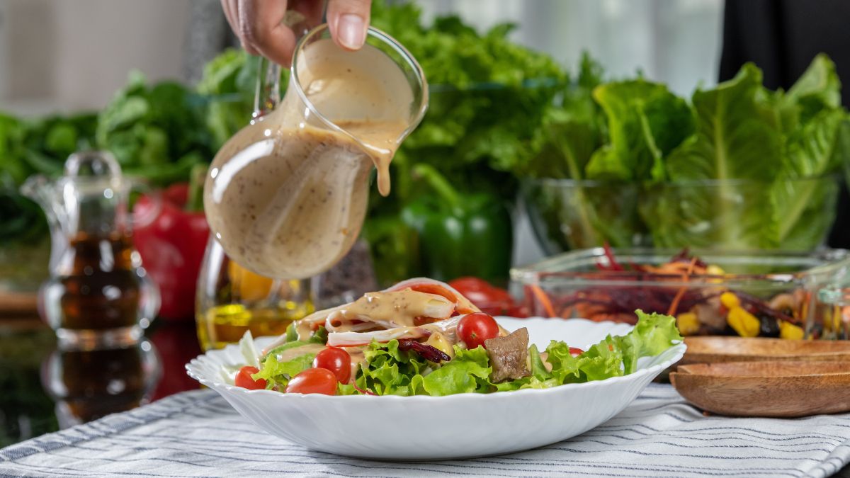 Cinco aderezos caseros en frascos de vidrio sobre mesa con ingredientes frescos: aguacate, miel, tahini, limones y hierbas aromáticas para ensaladas vibrantes